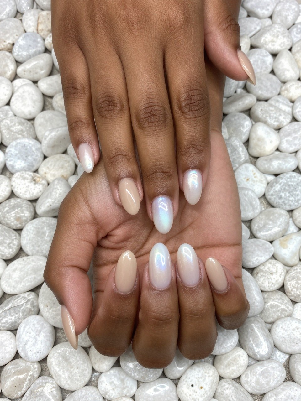 Top-down view of black feminine hands resting on a bed of smooth, white pebbles. The nails are a medium almond shape. The gradient goes from a matte sand beige at the cuticles to a pearly, iridescent shell-white at the tips. The light shows off the pearl finish.