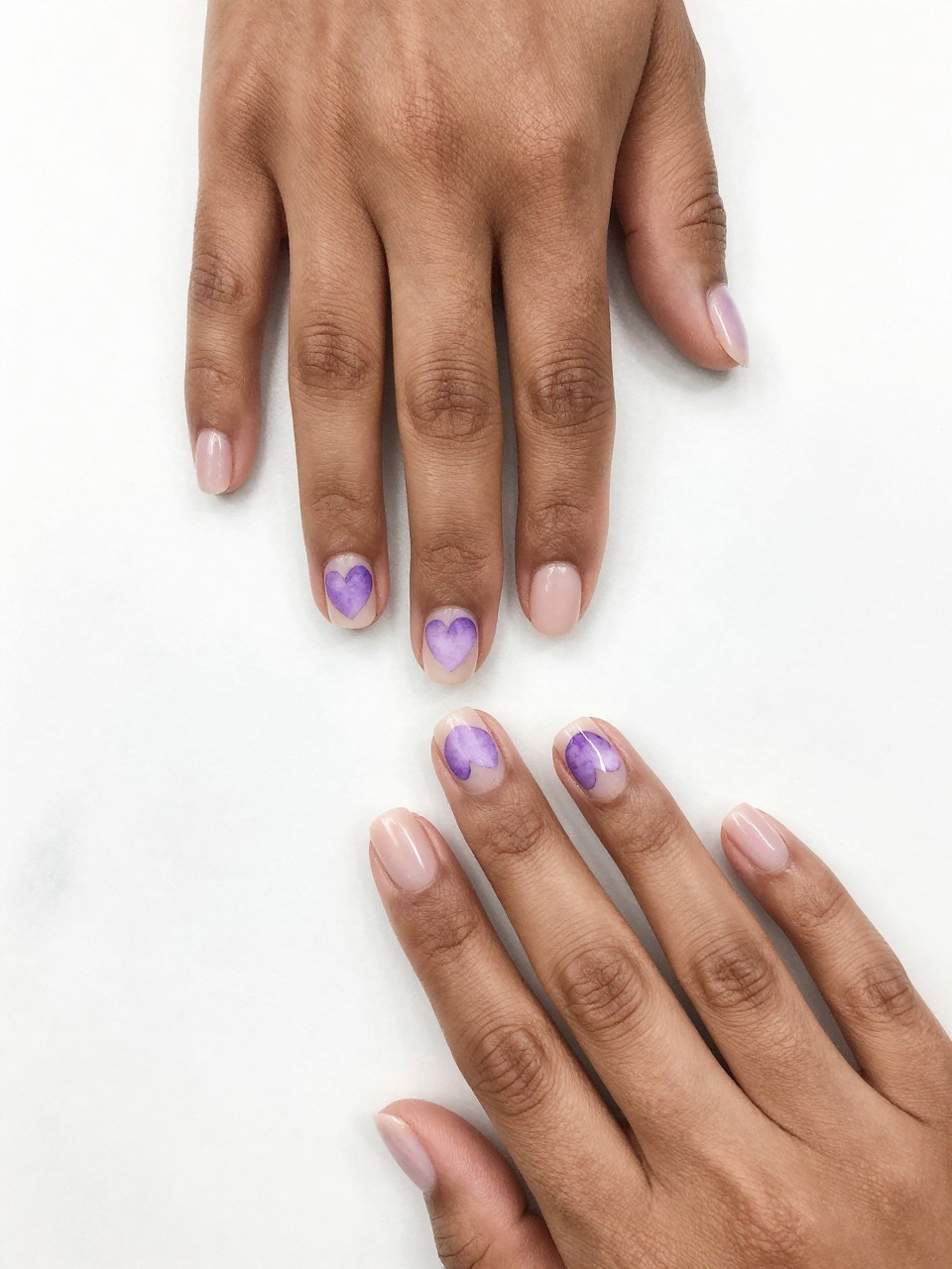 Overhead view of black feminine hands with clean, natural nails on a white marble surface. Each nail features a single, soft-focus watercolor heart in lavender hues. The look is minimalist, airy, and creatively gentle.