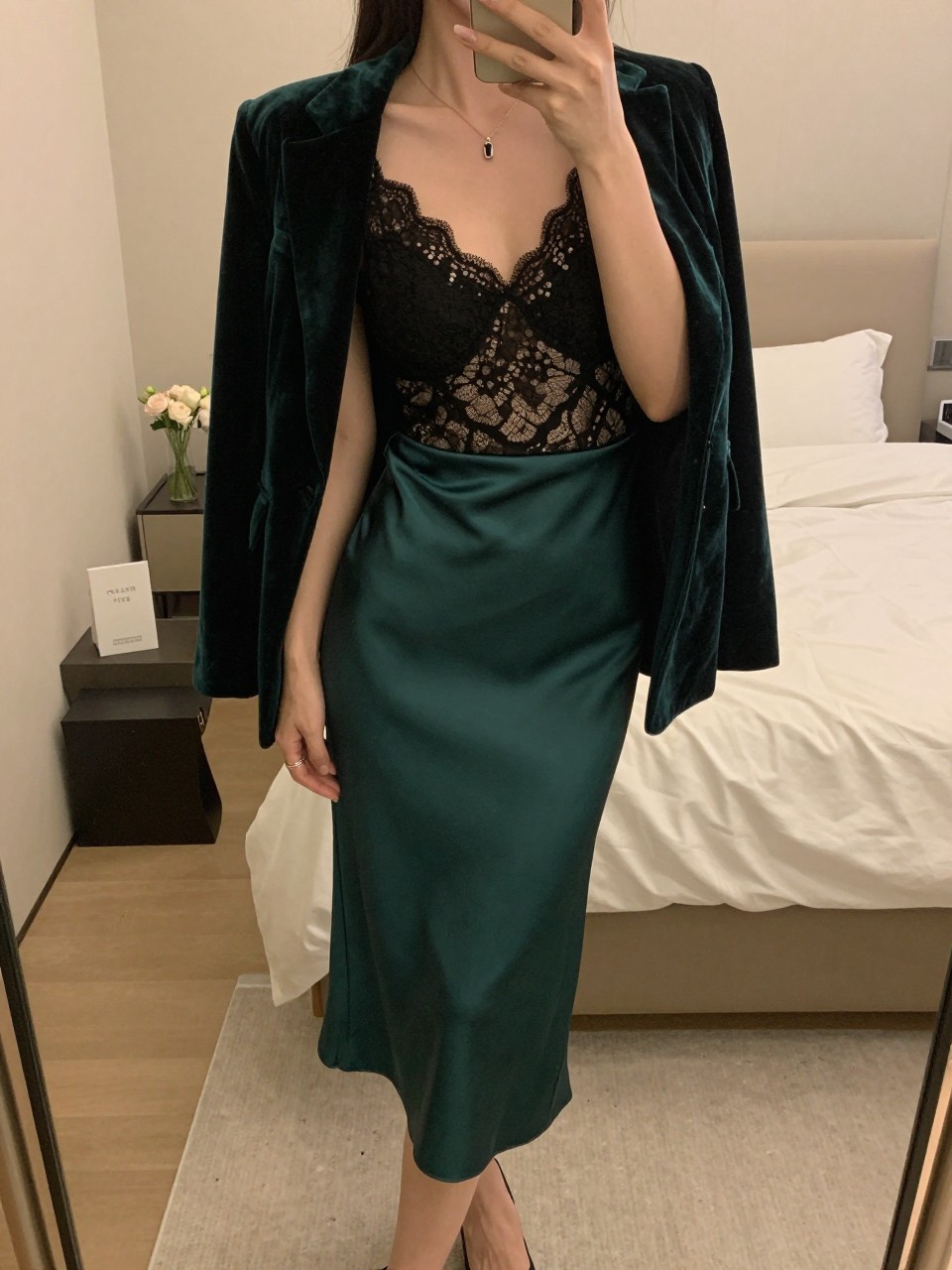 iPhone mirror selfie of a woman wearing a black lace cami with an emerald velvet blazer open over it, tucked into an emerald satin midi skirt with black pumps, one hand adjusting her blazer, cropped at NECK so face doesn't show, in a feminine-themed bedroom, warm moody lighting, iPhone photo quality.