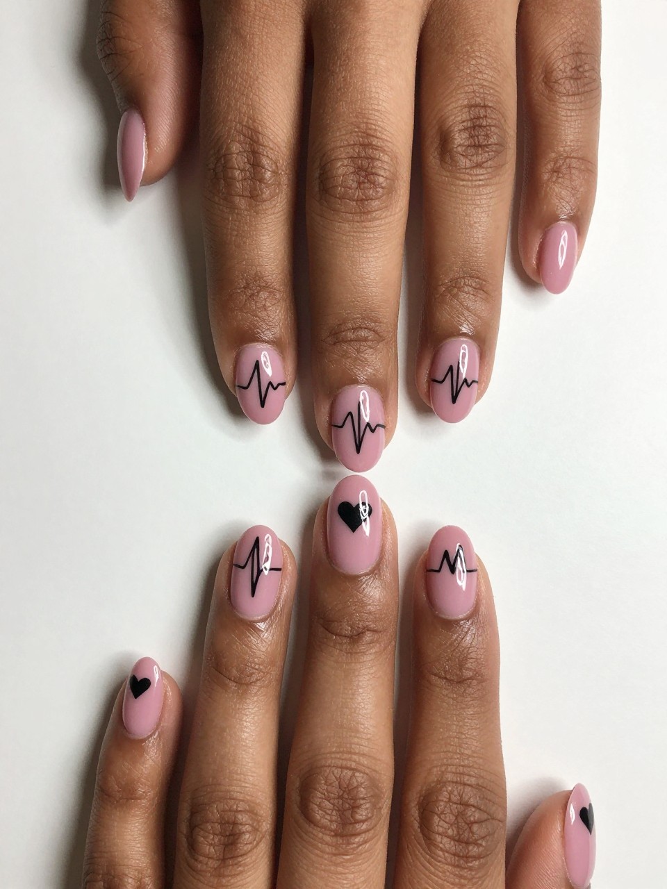Overhead shot of black feminine hands with oval nails on a plain background. The nails are a solid dusty pink. A single, wavy black heartbeat line runs across each nail, culminating in a small heart at the tip. The design is modern, meaningful, and subtly adorable.