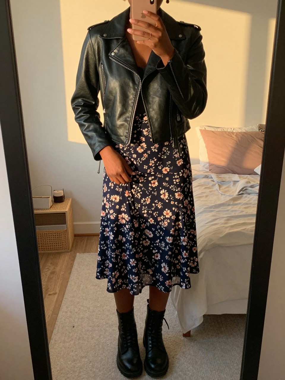 iPhone mirror selfie of a black woman in a cropped black leather jacket and a dark floral skirt with combat boots, one hand in her jacket pocket, cropped at NECK so face doesn't show, in a feminine-themed bedroom, evening golden hour, iPhone photo quality.