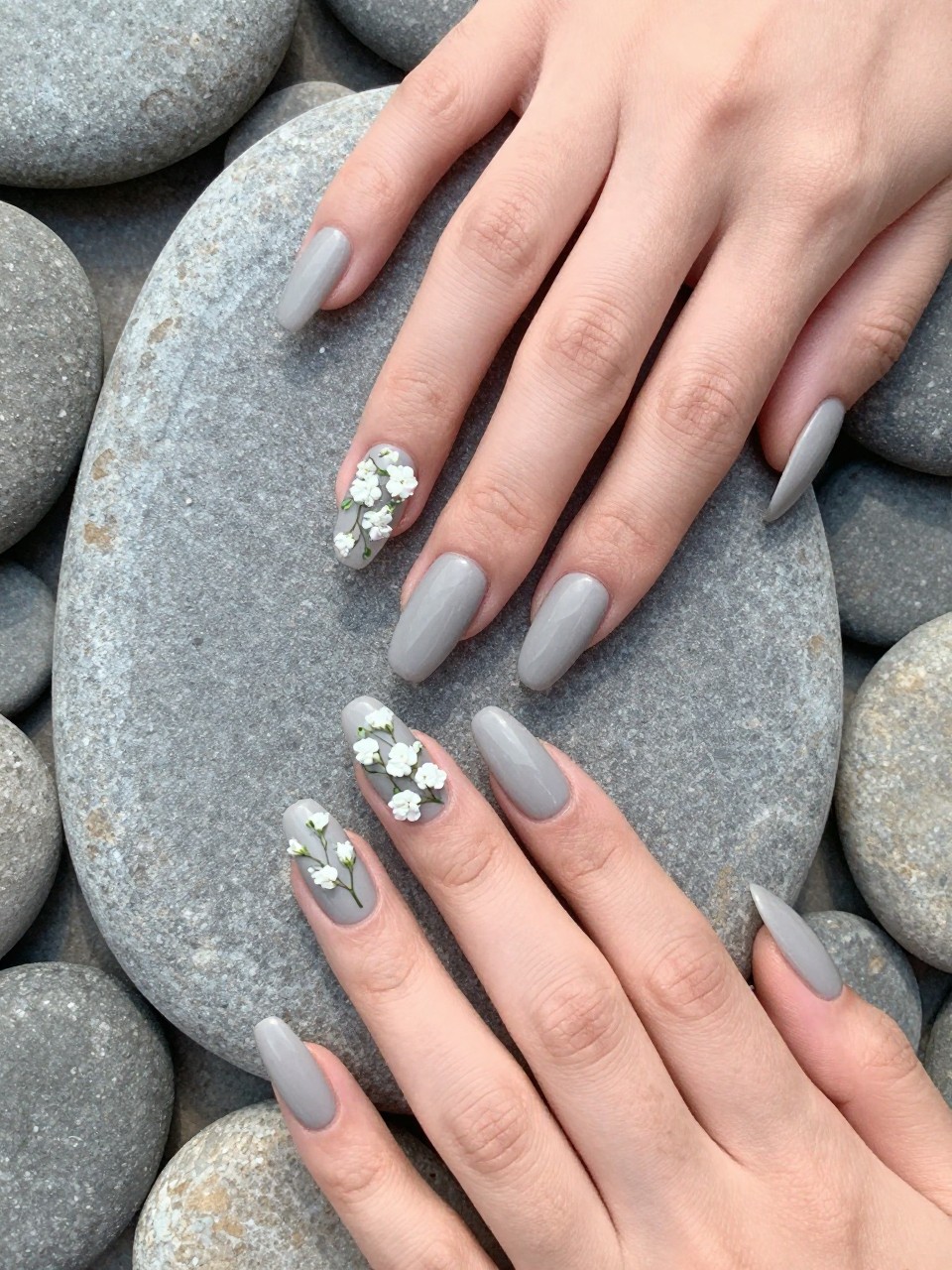 Bird's-eye view of hands with long nails resting on a smooth grey river stone. The nails are a matte stone grey. Delicate clusters of white baby’s breath flowers with micro green stems are painted on each nail. The mood is calm, clean, and fresh.