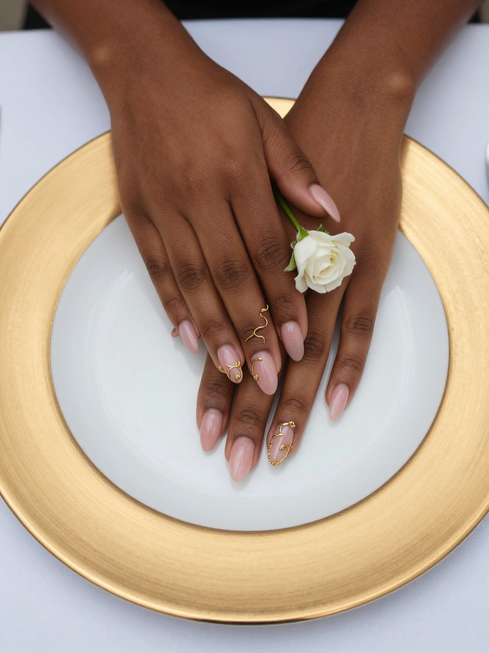 Overhead view of black feminine hands with almond nails resting on a gold-rimmed china plate at the reception table. A delicate gold vine curls from cuticle to tip on a sheer pink base, ending in a small white rose. The look is opulent and precise.