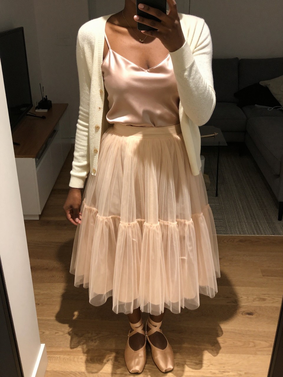 mirror selfie of a black woman wearing a blush silk camisole under a soft cream cardigan with a layered pastel tulle fairy skirt and nude ballet flats, one hand holding her phone at her side, cropped at NECK so face doesn't show, in a city apartment, warm moody lighting, iPhone photo quality.