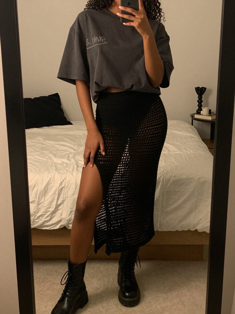 iPhone mirror selfie of a black woman wearing an oversized band tee with a black crochet midi skirt and black combat boots, one hand holding her phone low by her thigh, cropped at NECK so face doesn't show, in a feminine-themed bedroom, warm moody lighting, iPhone photo quality.