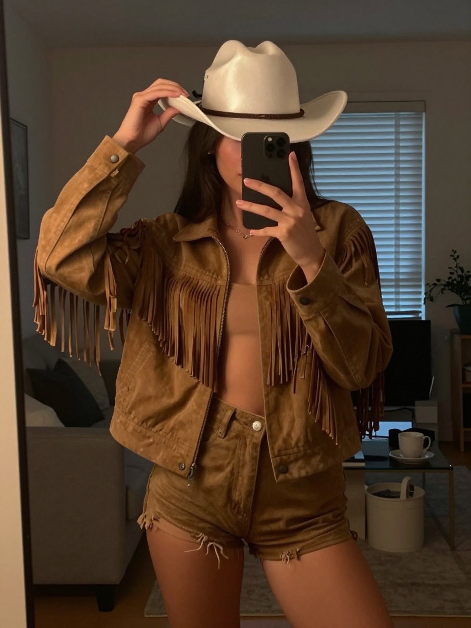 mirror selfie of a woman in a fringe jacket and shorts, adjusting a satin cowboy hat, cropped at NECK so face doesn't show, in a city apartment, warm moody lighting, iPhone photo quality.