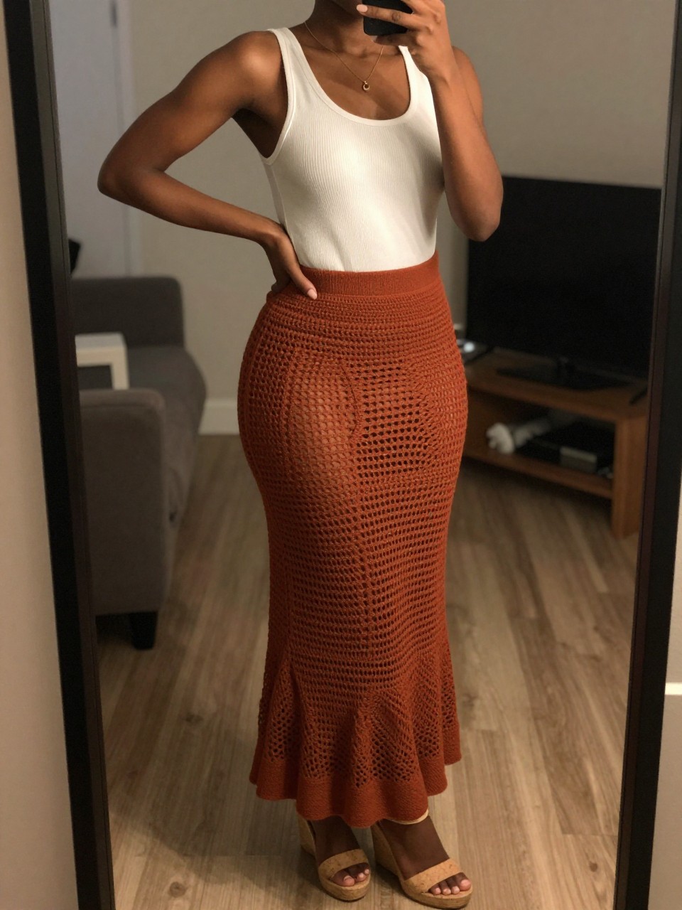 iPhone mirror selfie of a black woman wearing a white ribbed bodysuit tucked into a rust-colored crochet maxi skirt with natural cork wedge sandals, one hand on her hip, cropped at NECK so face doesn't show, in a city apartment, warm moody lighting, iPhone photo quality.