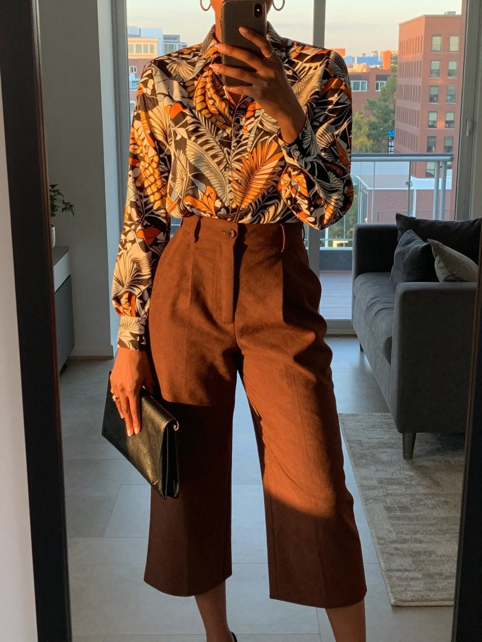 mirror selfie of a black woman in a printed blouse and culottes, holding a clutch in front, cropped at NECK so face doesn't show, in a city apartment, evening golden hour, iPhone photo quality.