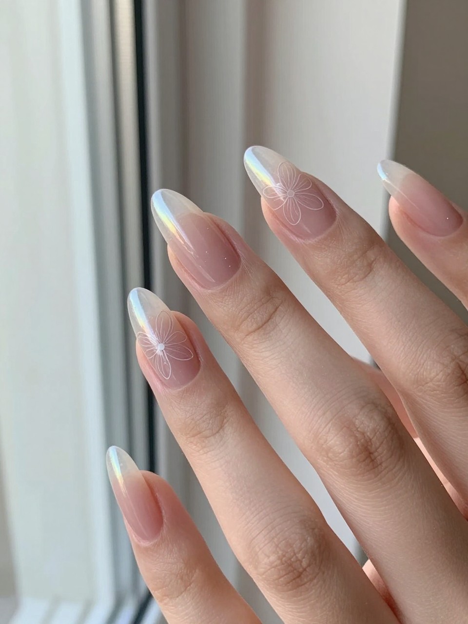 Hands with almond nails angled toward a window. The nails have a sheer opalescent base. A faint, same-toned flower outline is visible on each nail when the light hits it. The effect is magical, subtle, and luminous.