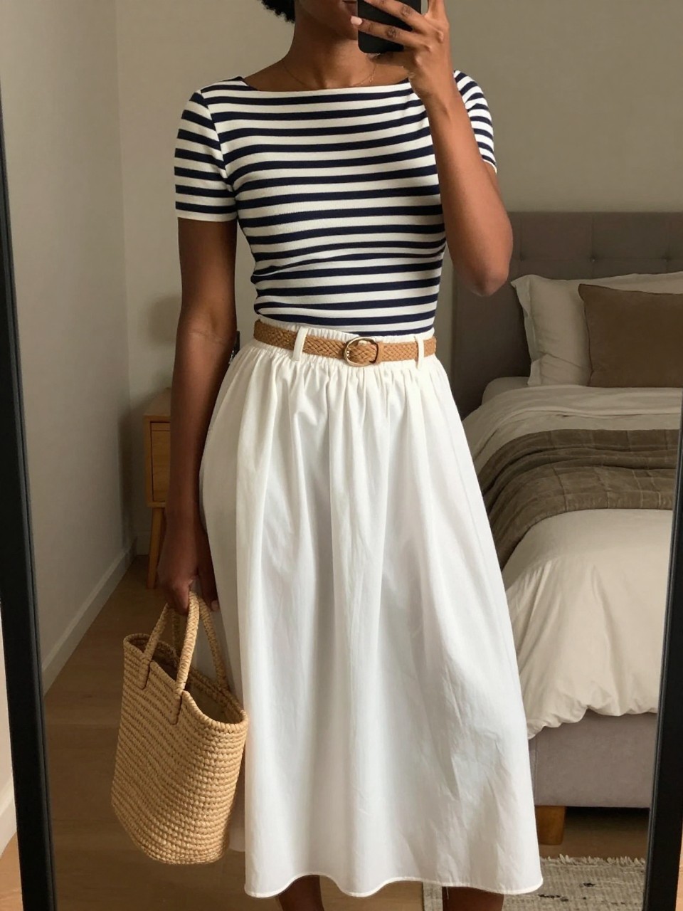 mirror selfie of a black woman wearing a fitted navy and white striped boatneck top tucked into a flowy white midi skirt with a tan woven belt cinching her waist, holding a straw tote bag, standing in a relaxed pose looking slightly away from the mirror, cropped at NECK so face doesn't show, in a feminine-themed bedroom, warm moody lighting, iPhone photo quality.