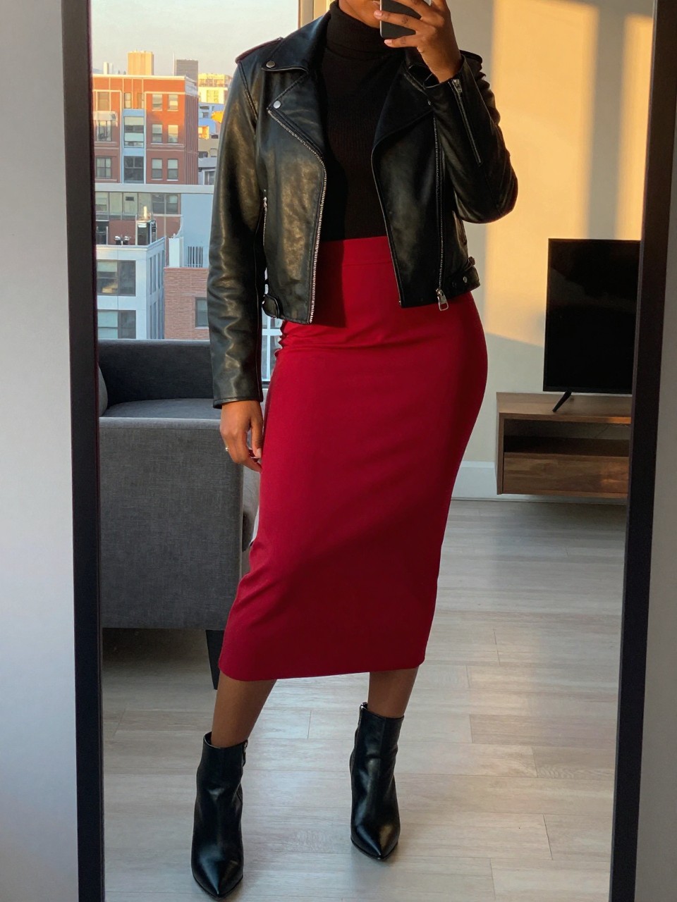 mirror selfie of a black woman wearing a black turtleneck tucked into a cherry red midi skirt with a black leather jacket over top and black heeled ankle boots, standing in a confident pose with one hand in her pocket, cropped at NECK so face doesn't show, in a city apartment, evening golden hour, iPhone photo quality.