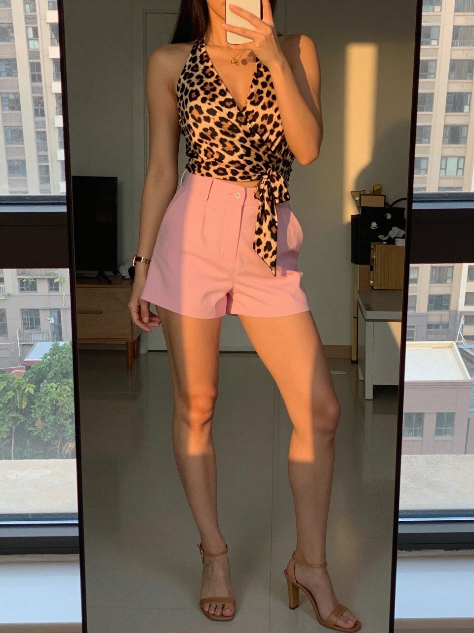 iPhone mirror selfie of a woman in pink shorts and a leopard wrap top with heeled sandals, standing with weight shifted, cropped at NECK so face doesn't show, in a city apartment, evening golden hour, iPhone photo quality.