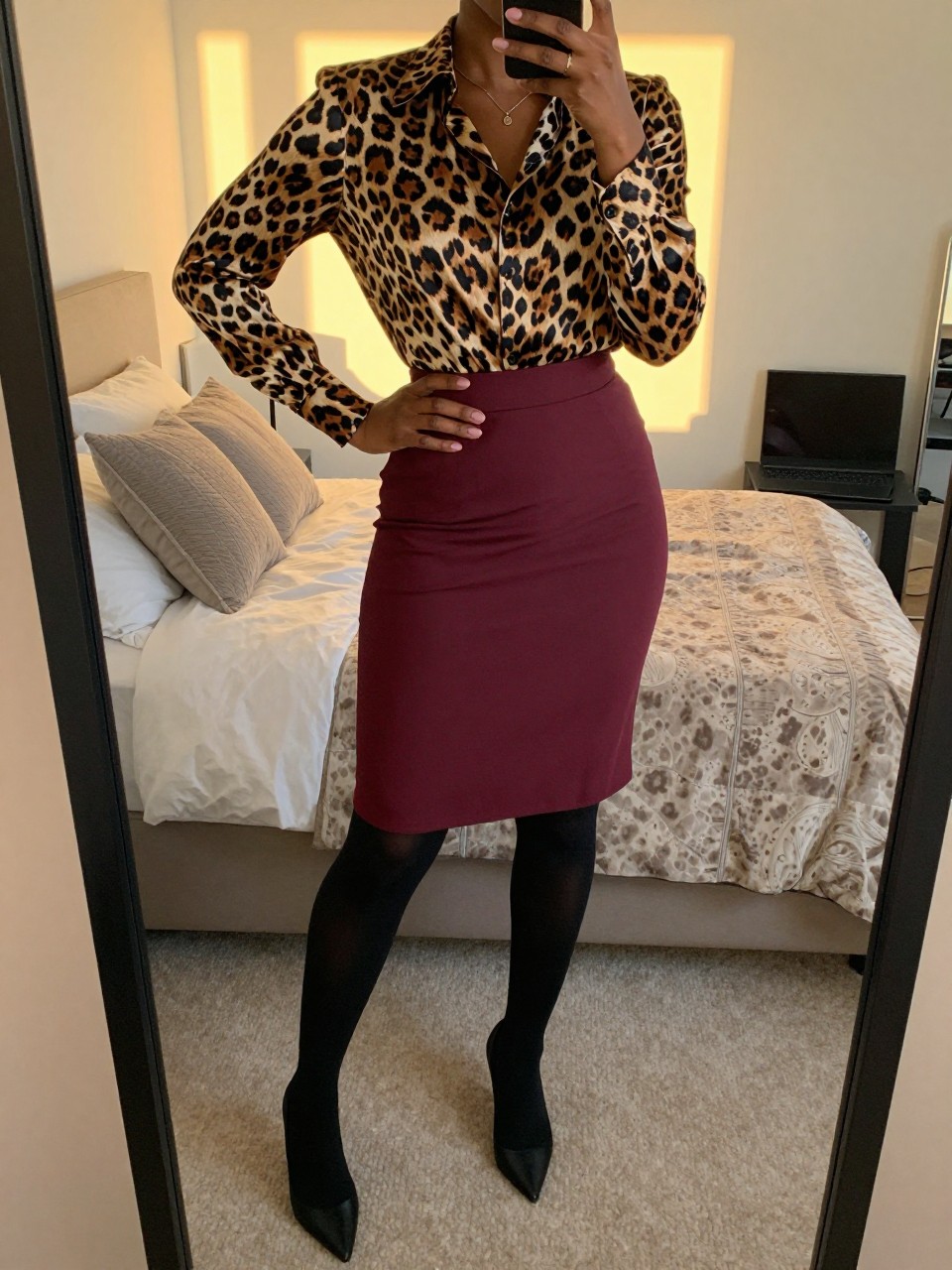 mirror selfie of a black woman wearing a leopard print silk blouse tucked into a maroon A-line skirt with black tights and pointed heels, one hand on her hip, cropped at NECK so face doesn't show, in a feminine-themed bedroom, evening golden hour, iPhone photo quality.
