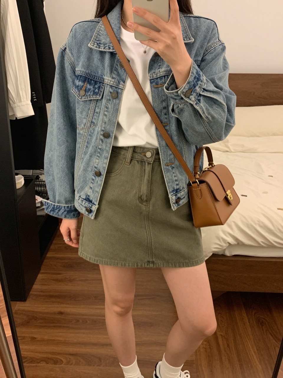 iPhone mirror selfie of a woman wearing a white tee tucked into an olive green denim mini skirt with a light-wash denim jacket worn open and white sneakers, holding a tan leather crossbody bag, standing in a casual weekend pose, cropped at NECK so face doesn't show, in a feminine-themed bedroom, warm moody lighting, iPhone photo quality.