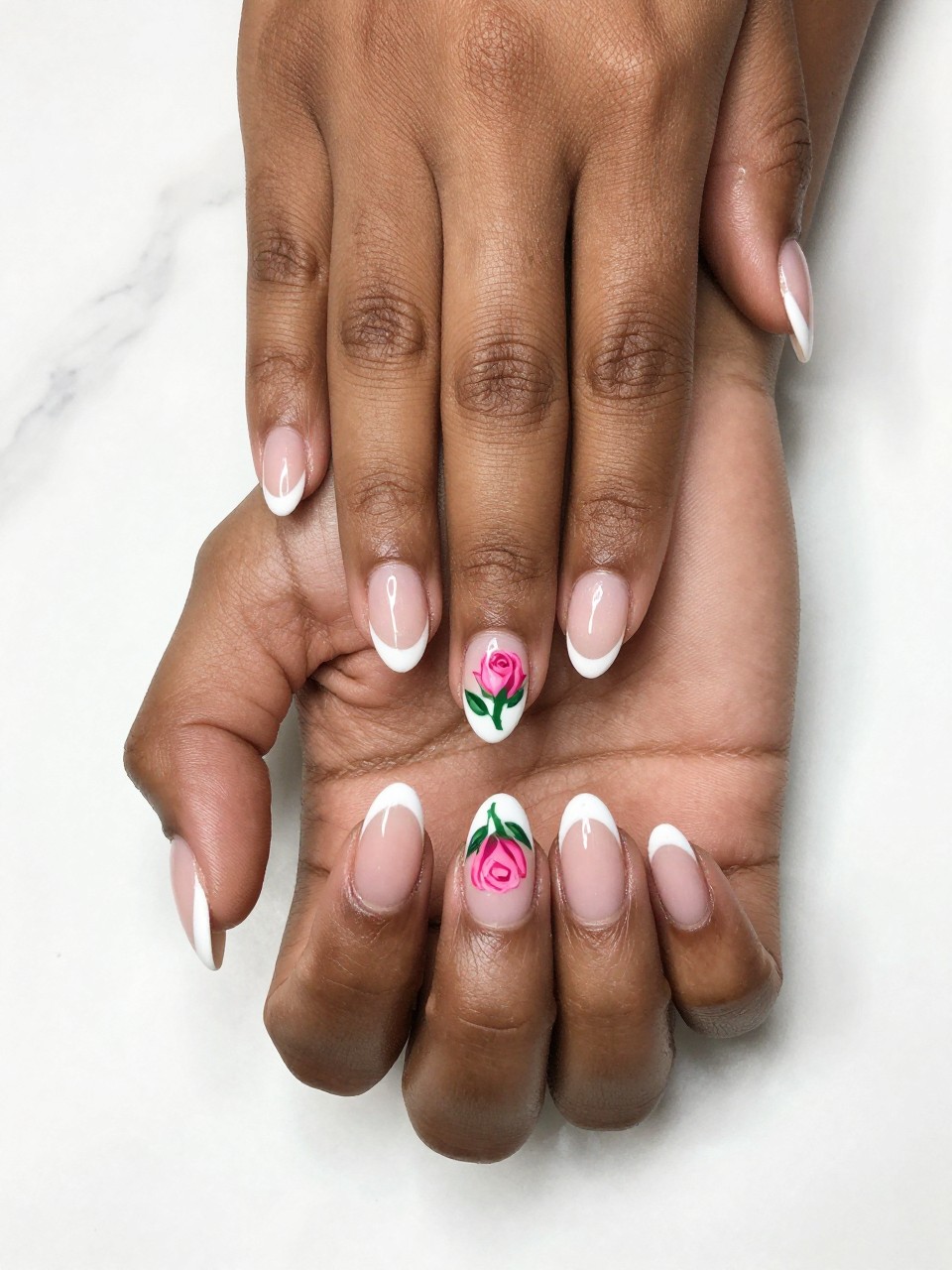 Overhead, top-down view of black feminine hands with medium almond nails resting on a white marble surface. The nails have a classic white French tip. A single, detailed pink rosebud with a green leaf is painted on the side of each nail bed. The lighting is bright and clean, showcasing the precise details.
