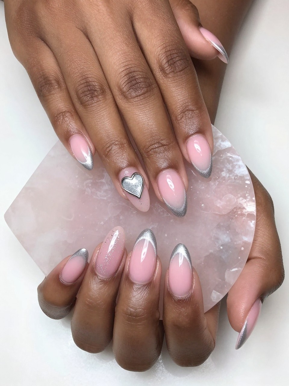 Bird's-eye view of black feminine hands with almond nails on a rose quartz slab. The nails show a pink ombré with a sweeping silver cat eye effect. One accent nail features a thin, chrome outline of a heart over the sparkle. The setting is soft and luminous.