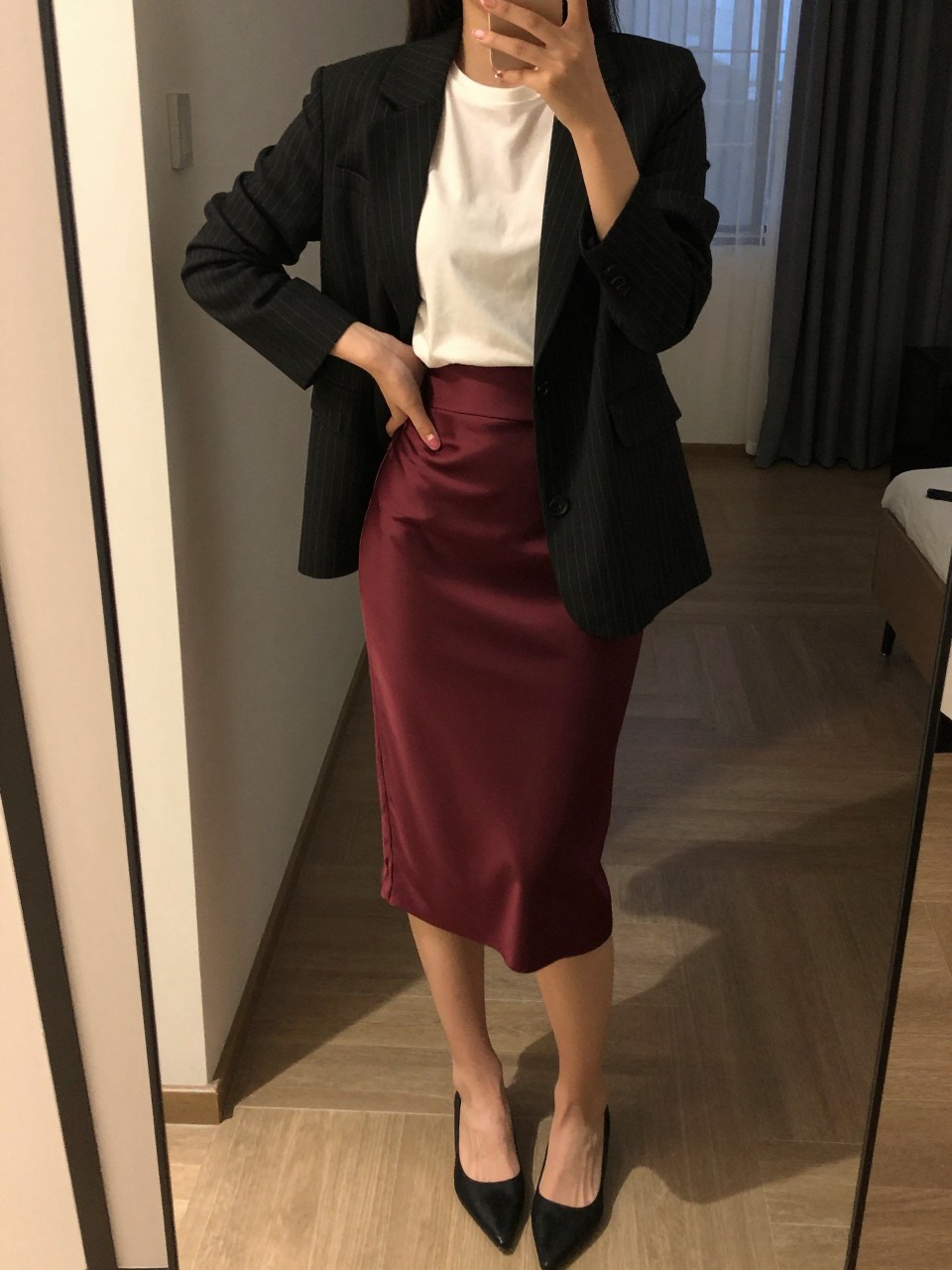 mirror selfie of a woman wearing a black pinstripe blazer over a white tee tucked into a maroon satin pencil skirt with black pointed flats, holding her phone by her hip, cropped at NECK so face doesn't show, in a city apartment, warm moody lighting, iPhone photo quality.