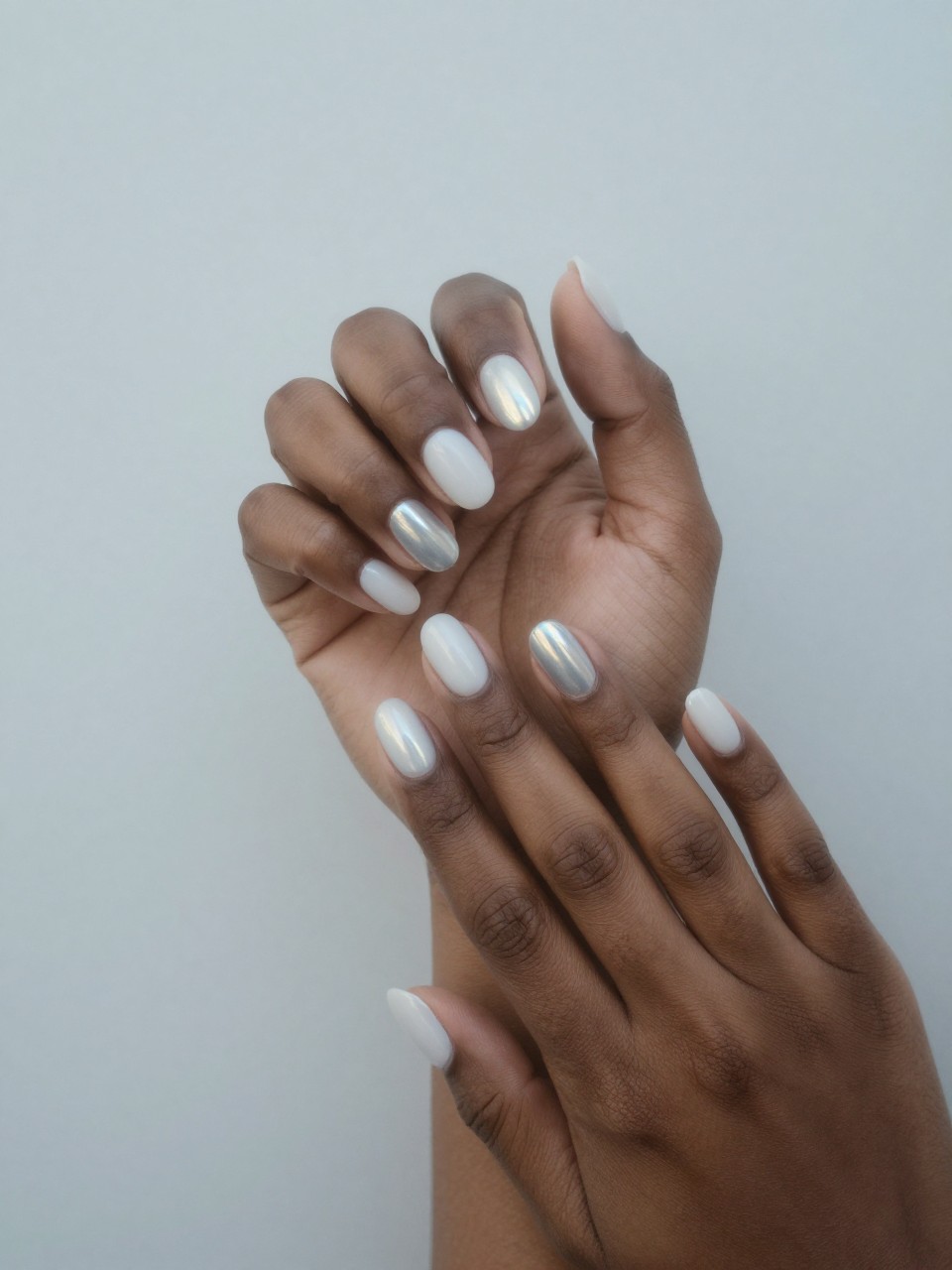Overhead view of black feminine hands against a misty, light grey backdrop. The nails are oval with a gradient from a soft matte white to a smooth, holographic silver polish at the tips that reflects rainbow colors. Diffused, ethereal lighting completes the scene.