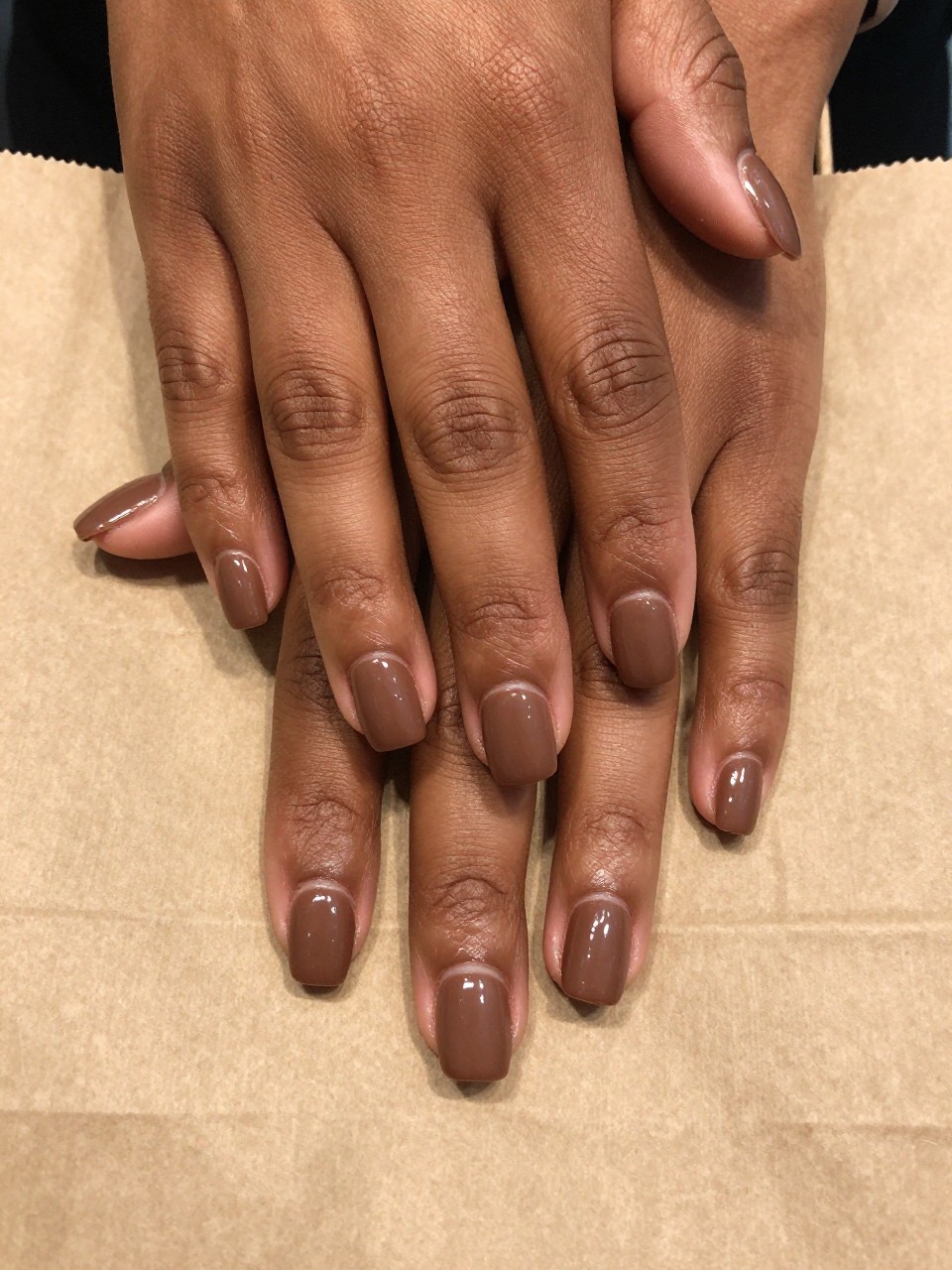 Close-up, top-down view of black feminine hands with short, square nails resting on a kraft paper bag. The nails are a warm, matte chocolate brown. The lunula at the base of each nail is shiny and glossy, creating a subtle two-tone effect. The lighting is even and clear.