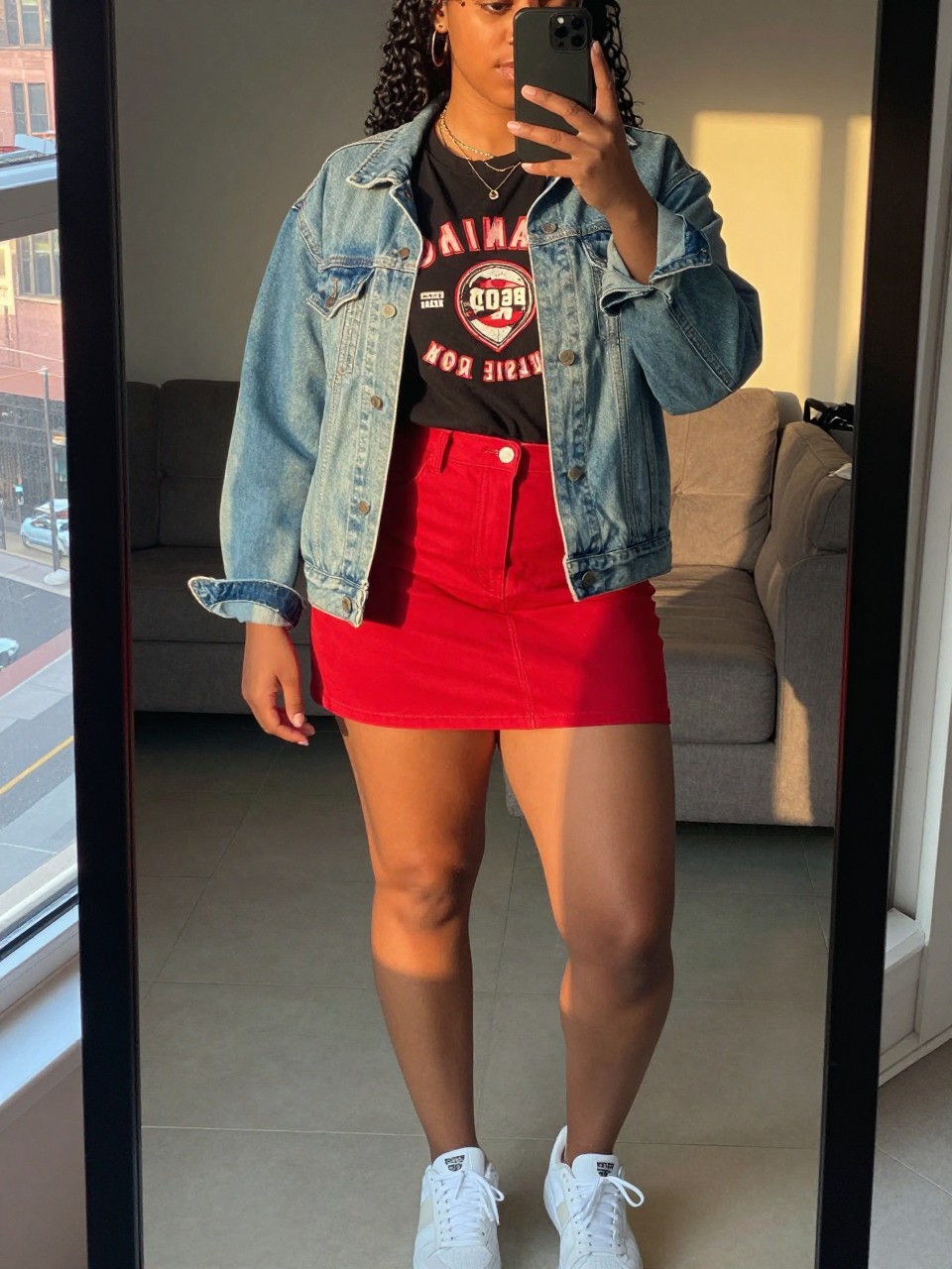 mirror selfie of a curvy black woman wearing a black vintage band tee tucked into a cherry red denim mini skirt with a light-wash denim jacket worn open and white sneakers, sunglasses perched on her head, standing in a casual weekend pose, cropped at NECK so face doesn't show, in a city apartment, evening golden hour, iPhone photo quality.