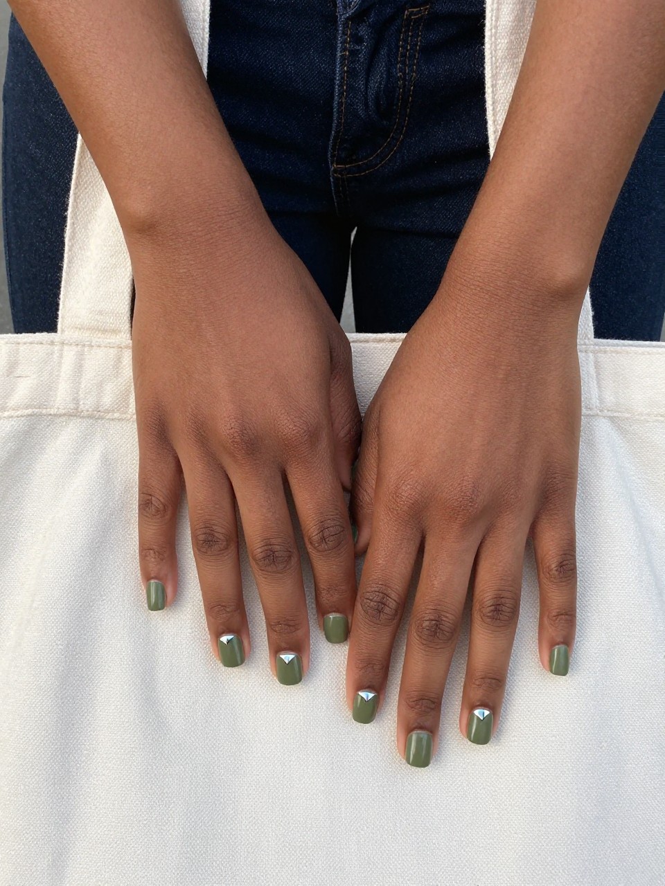 Overhead view of black feminine hands with short, squared nails lightly gripping the handle of a canvas tote bag. The nails are a matte olive green. A single, small silver pyramid stud sits at the base of each ring finger nail. The setting is casual and urban.