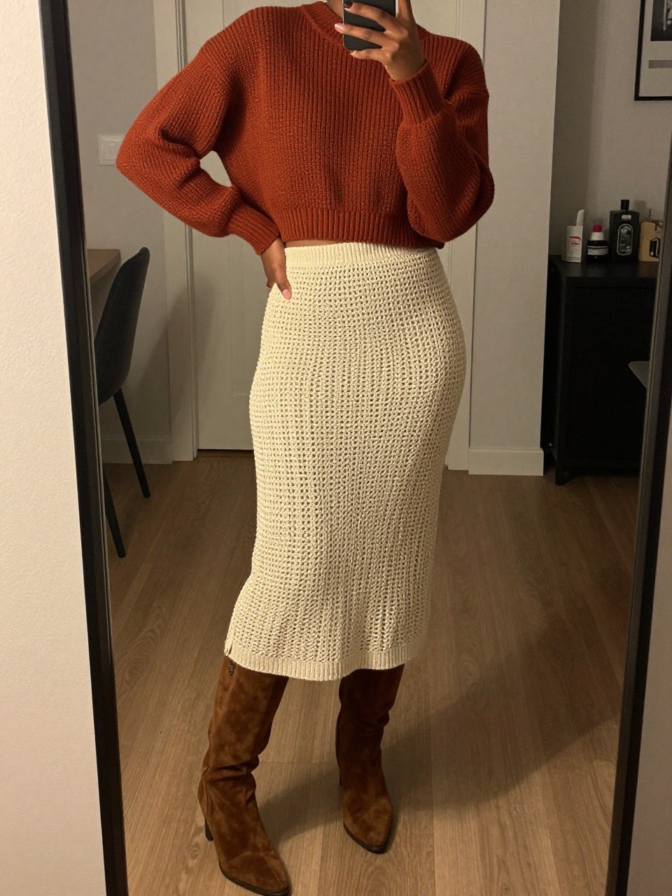 iPhone mirror selfie of a black woman wearing a rust cropped ribbed sweater with a natural crochet midi skirt and brown suede knee-high boots, one hand on her hip, cropped at NECK so face doesn't show, in a city apartment, warm moody lighting, iPhone photo quality.
