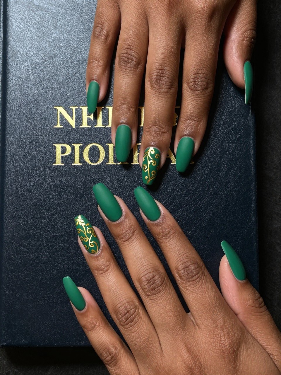 Overhead view of black feminine hands with long nails resting on a leather-bound book with gold foil lettering. The nails are a matte hunter green. An intricate, hand-painted gold vine pattern curls around each nail from cuticle to tip. The look is luxurious and detailed.