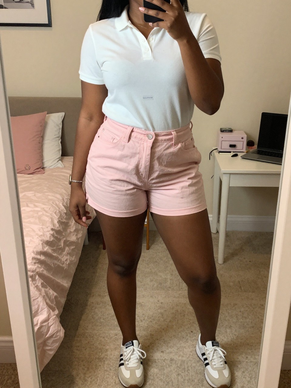 iPhone mirror selfie of a black curvy woman in pink chino shorts and a white polo with sneakers, standing with phone near waist, cropped at NECK so face doesn't show, in a feminine-themed bedroom, warm moody lighting, iPhone photo quality.