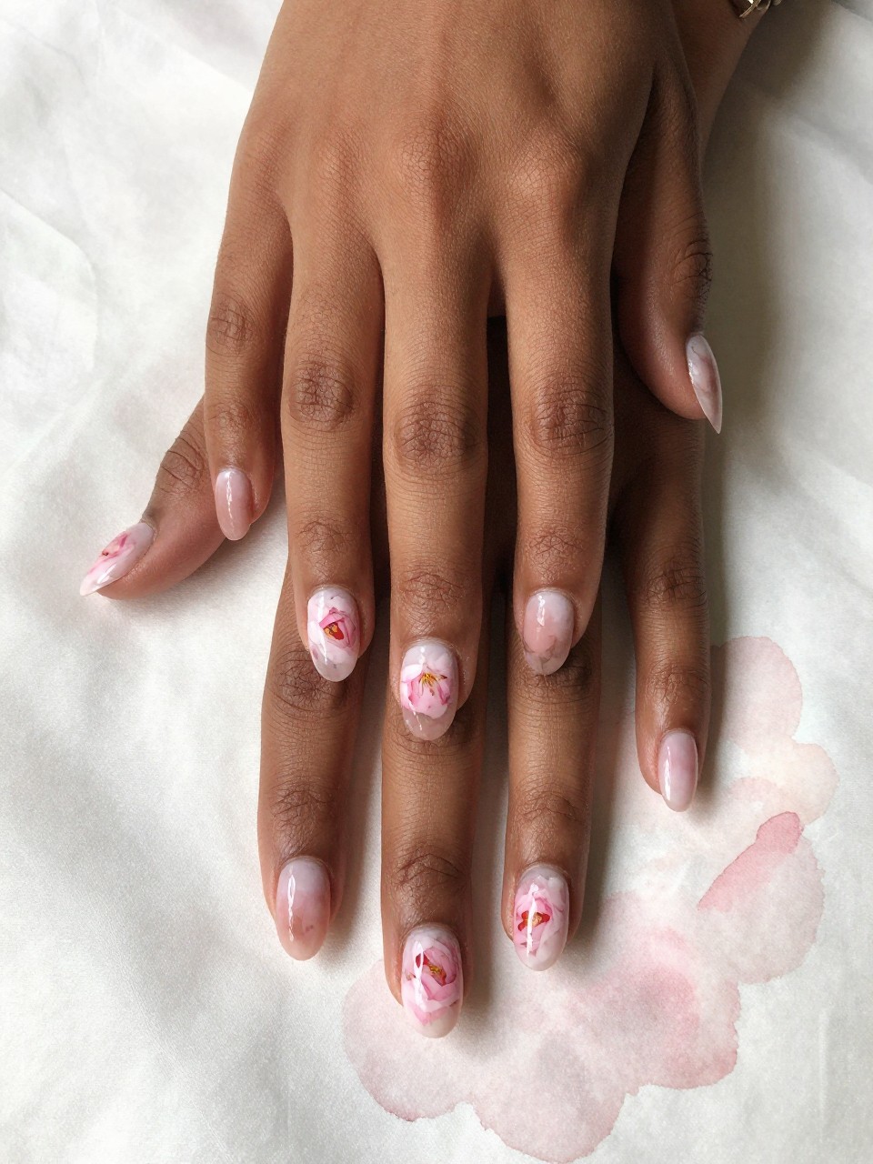 Top-down view of black feminine hands with oval nails on a silk watercolor painter’s cloth. Each nail has a soft, blurred wash of pale pink forming an abstract peony bloom. The style is impressionistic, romantic, and deeply artistic.