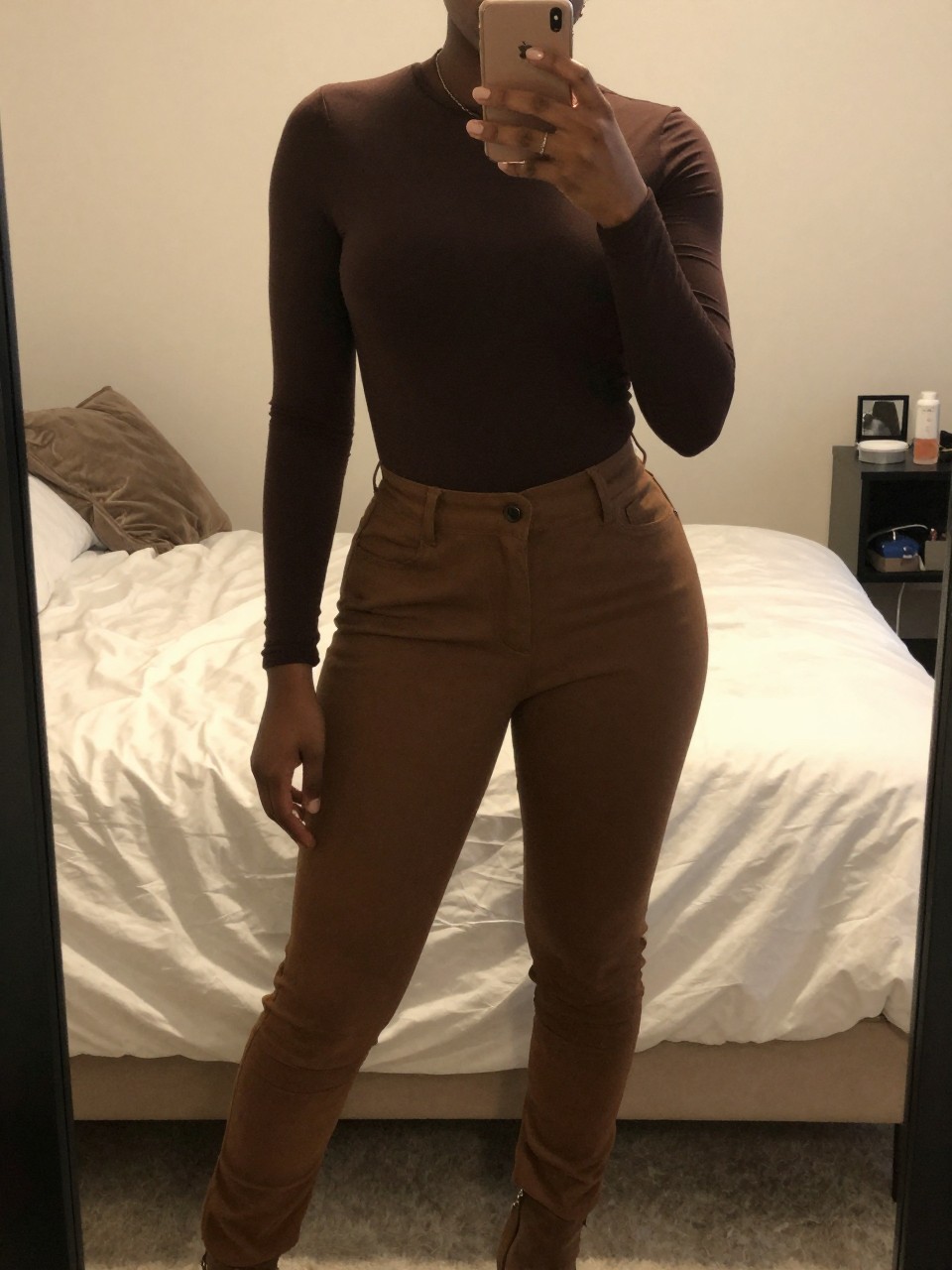 mirror selfie of a Black woman wearing a chocolate brown top with brown trousers and boots, posed with her weight shifted and phone held casually, cropped at NECK so face doesn't show, in a feminine-themed bedroom, warm moody lighting, iPhone photo quality.
