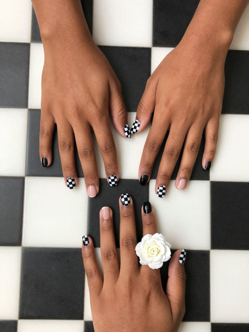 Top-down view of black feminine hands with short nails on a checkerboard floor tile. the nail designs are black and white checkered. The accent nails are solid black with one detailed white rose. The vibe is edgy, playful, and retro.