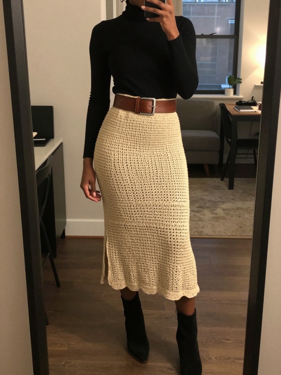 iPhone mirror selfie of a black woman wearing a black turtleneck tucked into a natural crochet midi skirt with a wide tooled leather belt and black suede ankle boots, one hand resting on the belt, cropped at NECK so face doesn't show, in a city apartment, warm moody lighting, iPhone photo quality.
