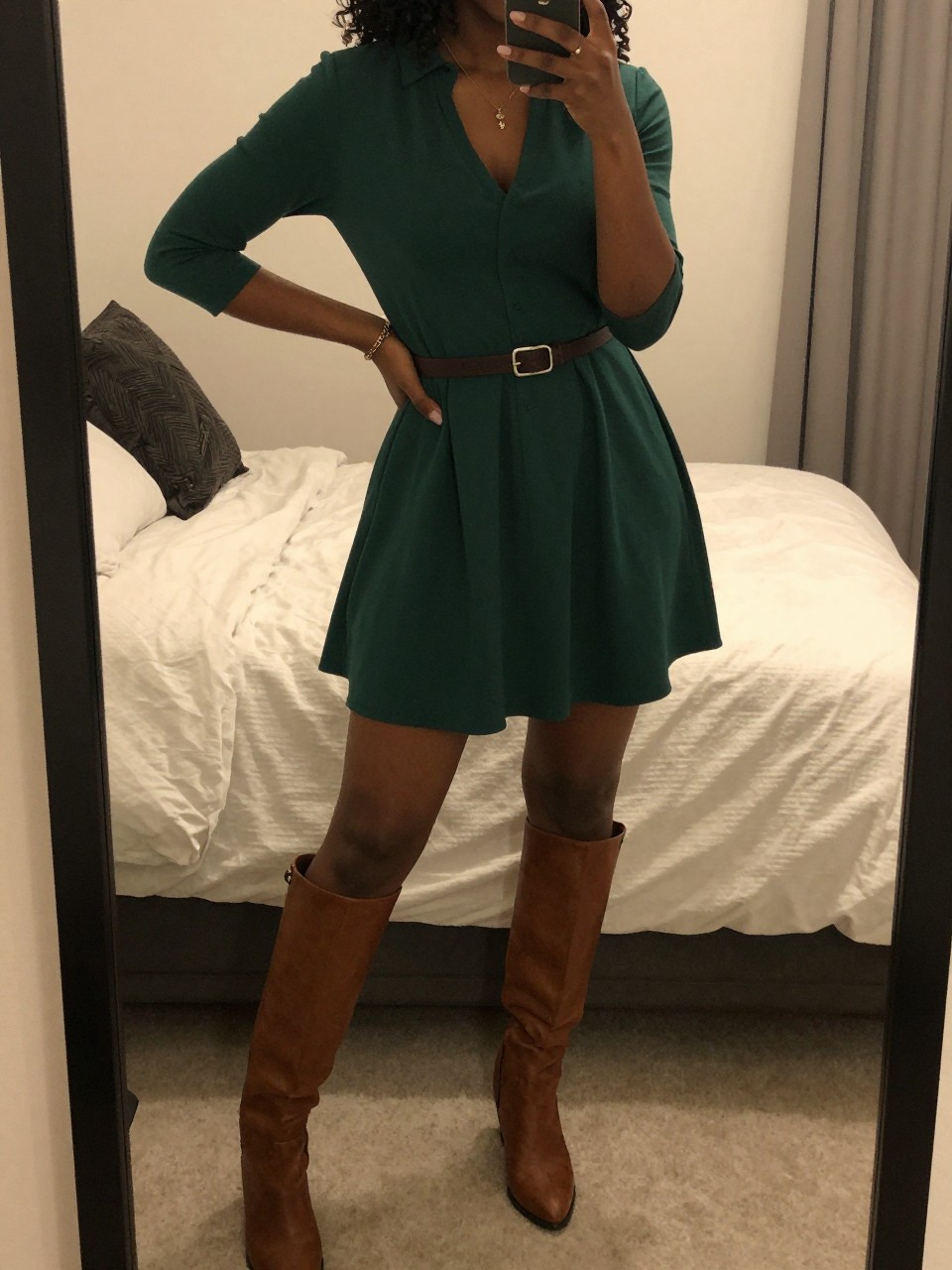 mirror selfie of a Black woman wearing a forest green dress with brown knee-high boots and a belt, posed with one hand on her hip and phone in the other, cropped at NECK so face doesn't show, in a feminine-themed bedroom, warm moody lighting, iPhone photo quality.