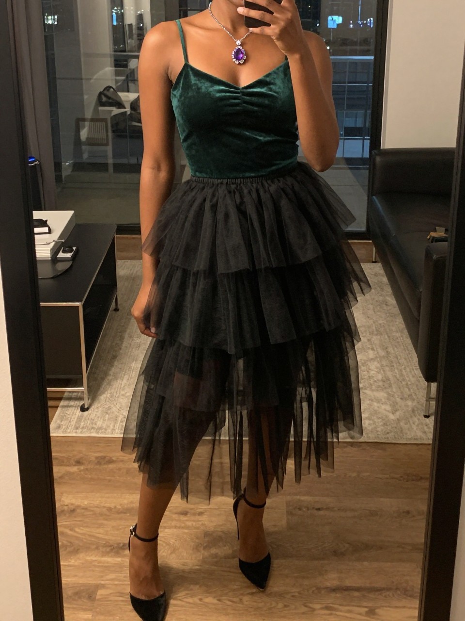 mirror selfie of a black woman wearing an emerald velvet camisole tucked into a black layered tulle fairy skirt with an amethyst crystal pendant necklace and black velvet heels, one hand touching her crystal, cropped at NECK so face doesn't show, in a city apartment, warm moody lighting, iPhone photo quality.