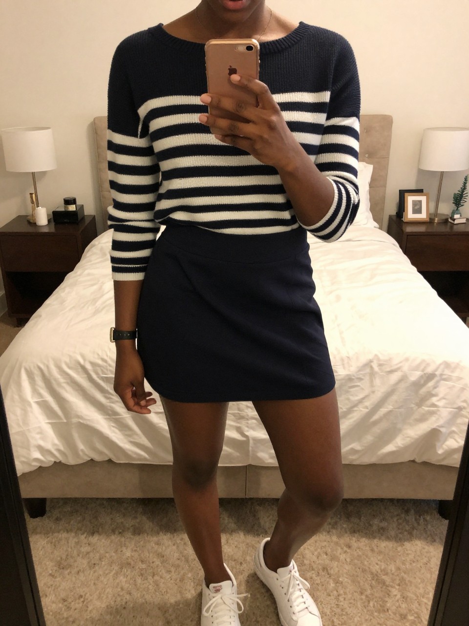 mirror selfie of a black woman wearing a navy striped boatneck sweater tucked into a navy tennis skirt with white sneakers, holding her phone at chest level, cropped at NECK so face doesn't show, in a feminine-themed bedroom, warm moody lighting, iPhone photo quality.