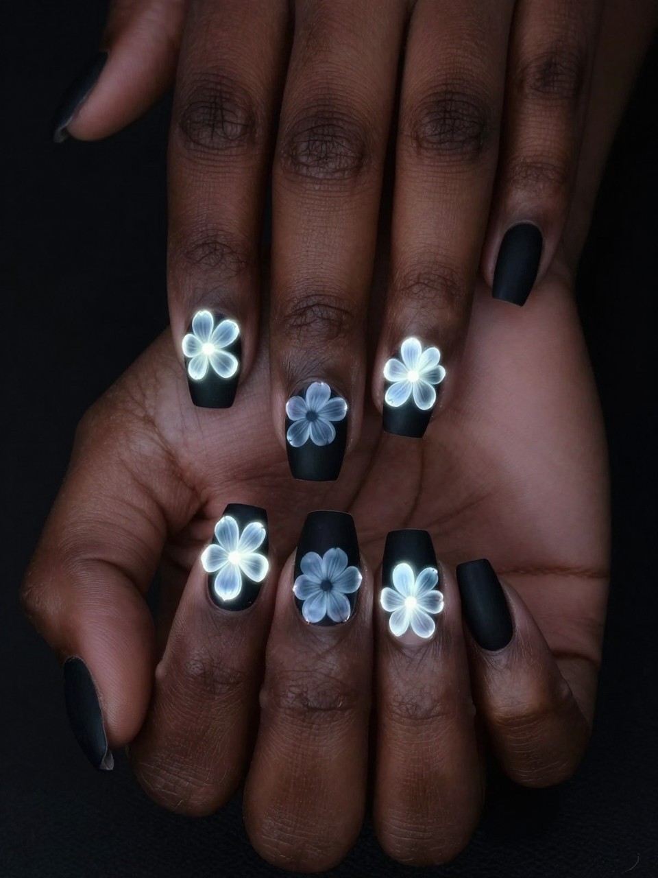 Close-up, top-down view of black feminine hands against a dark background. The nails are matte black. Luminous white flower outlines with soft, glowy edges are painted on each nail, as if they are bioluminescent. The mood is magical and otherworldly.