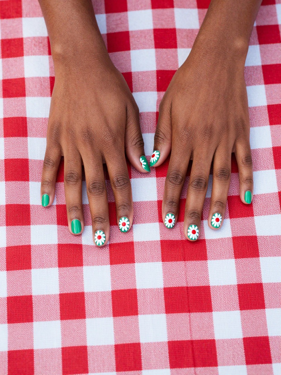Top-down view of black feminine hands with short nails on a red and white checkered picnic blanket. The nails are a glossy kelly green. Bold white daisies with bright red centers are painted on each nail. The scene is classic, fun, and full of vintage charm.