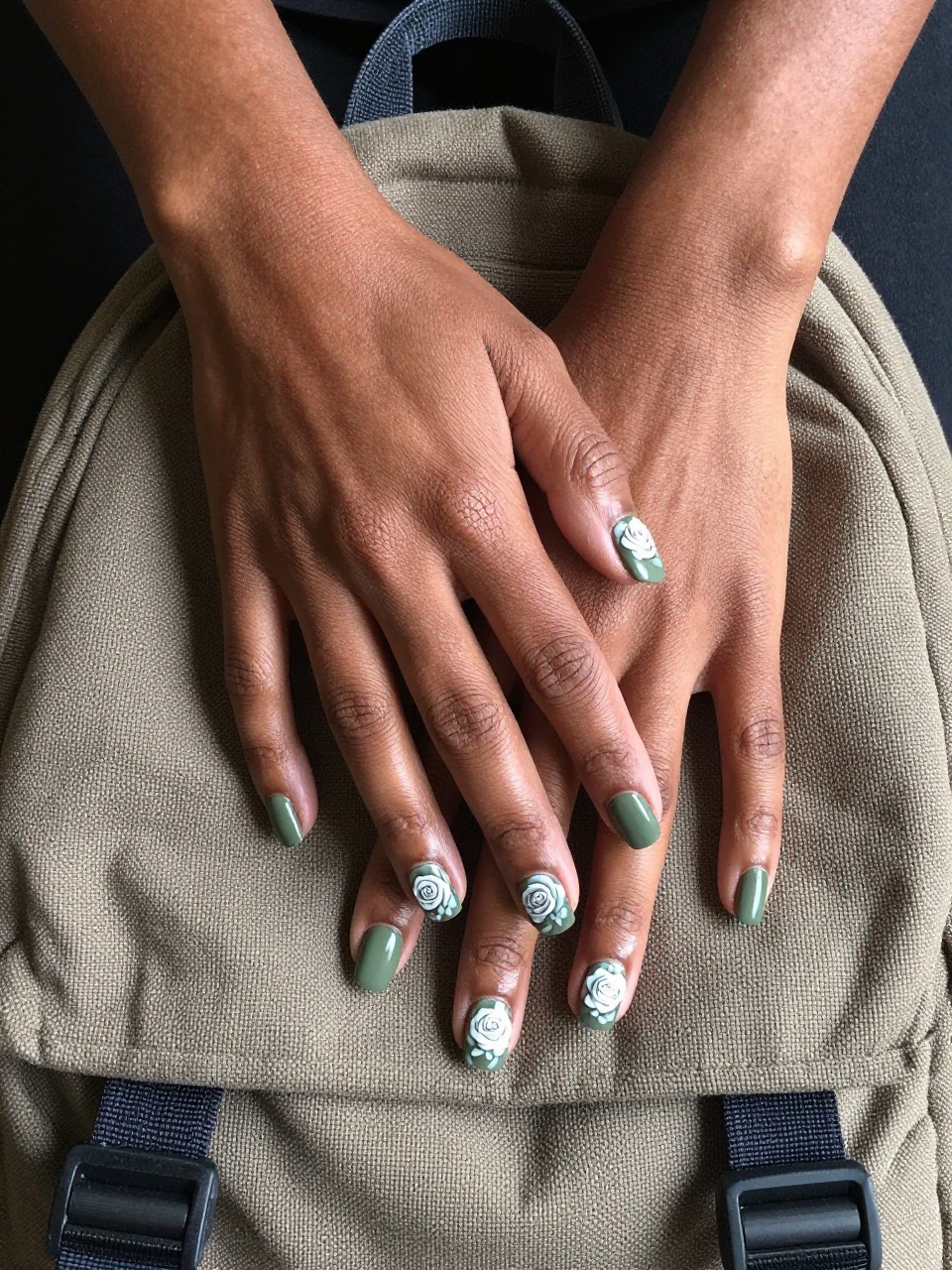 Overhead view of black feminine hands with short, squared nails resting on a canvas backpack. The nails are a matte army green and features single, beautifully painted white rose with light grey shading sits on each nail. The vibe is rugged yet romantic.