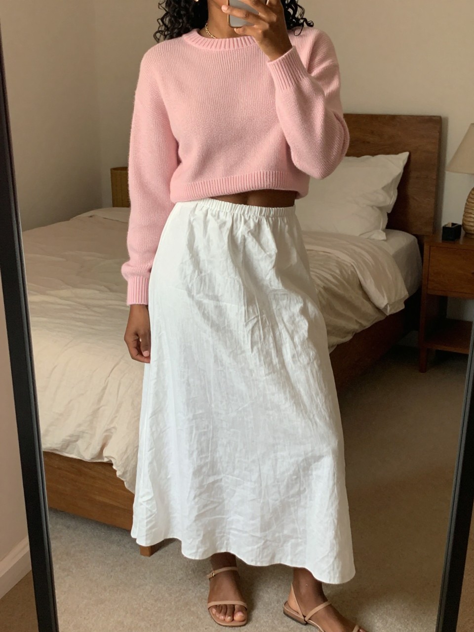 mirror selfie of a black woman wearing a pale pink cropped cashmere sweater with a white linen maxi skirt and thin nude flat sandals, a delicate anklet visible, standing in a relaxed pose, cropped at NECK so face doesn't show, in a feminine-themed bedroom, warm moody lighting, iPhone photo quality.