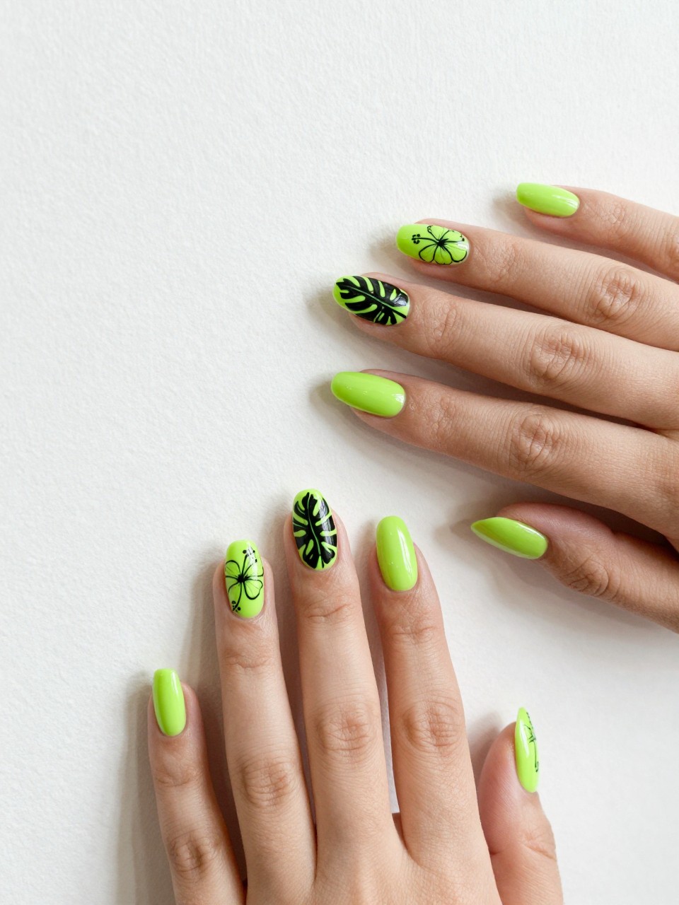 Overhead shot of hands with coffin nails design against a white minimalist wall. The nails are a glossy neon lime green. Bold, black outlined monstera leaves or hibiscus flowers are painted on each nail. The composition is sharp and contemporary.