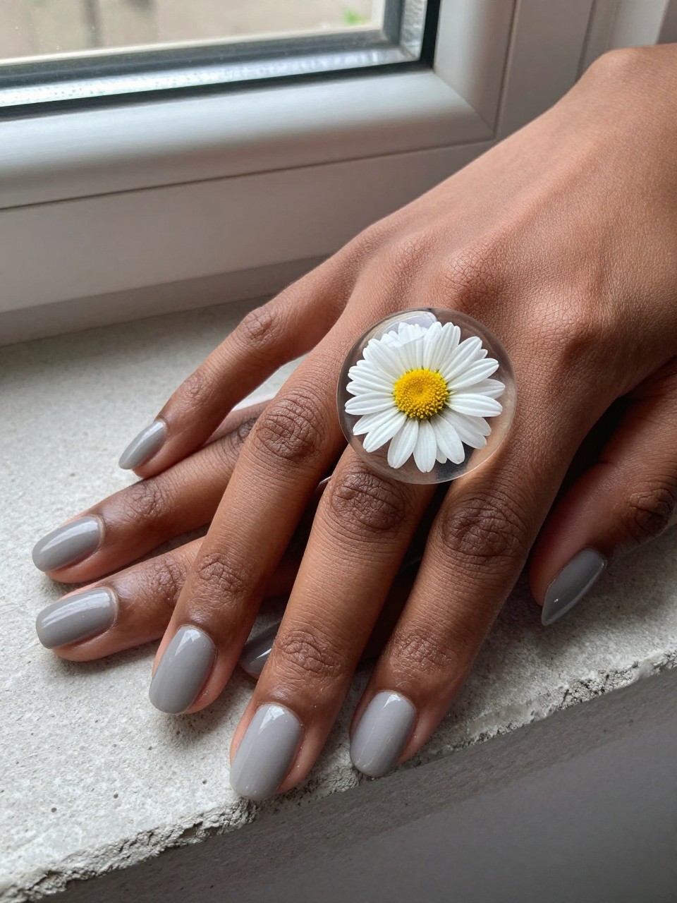 Macro, overhead view of black feminine hands with rounded nails over a concrete windowsill. The nails are a matte greige. The ring fingers have a single, real pressed white daisy perfectly preserved under a glossy dome. The look is modern yet organic.