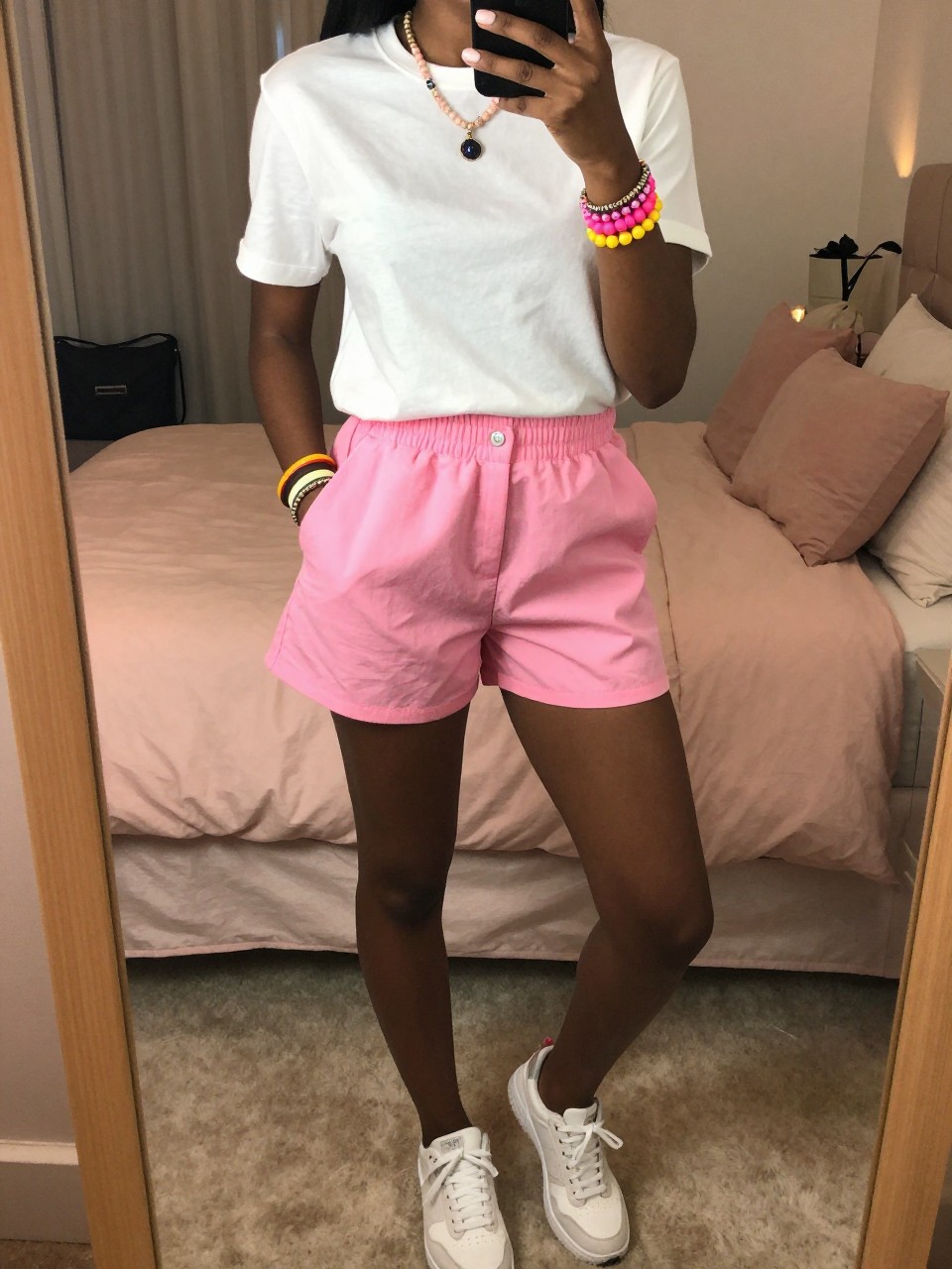 iPhone mirror selfie of a black woman in pink shorts and a white tee with colorful accessories and sneakers, standing with hand in pocket, cropped at NECK so face doesn't show, in a feminine-themed bedroom, warm moody lighting, iPhone photo quality.