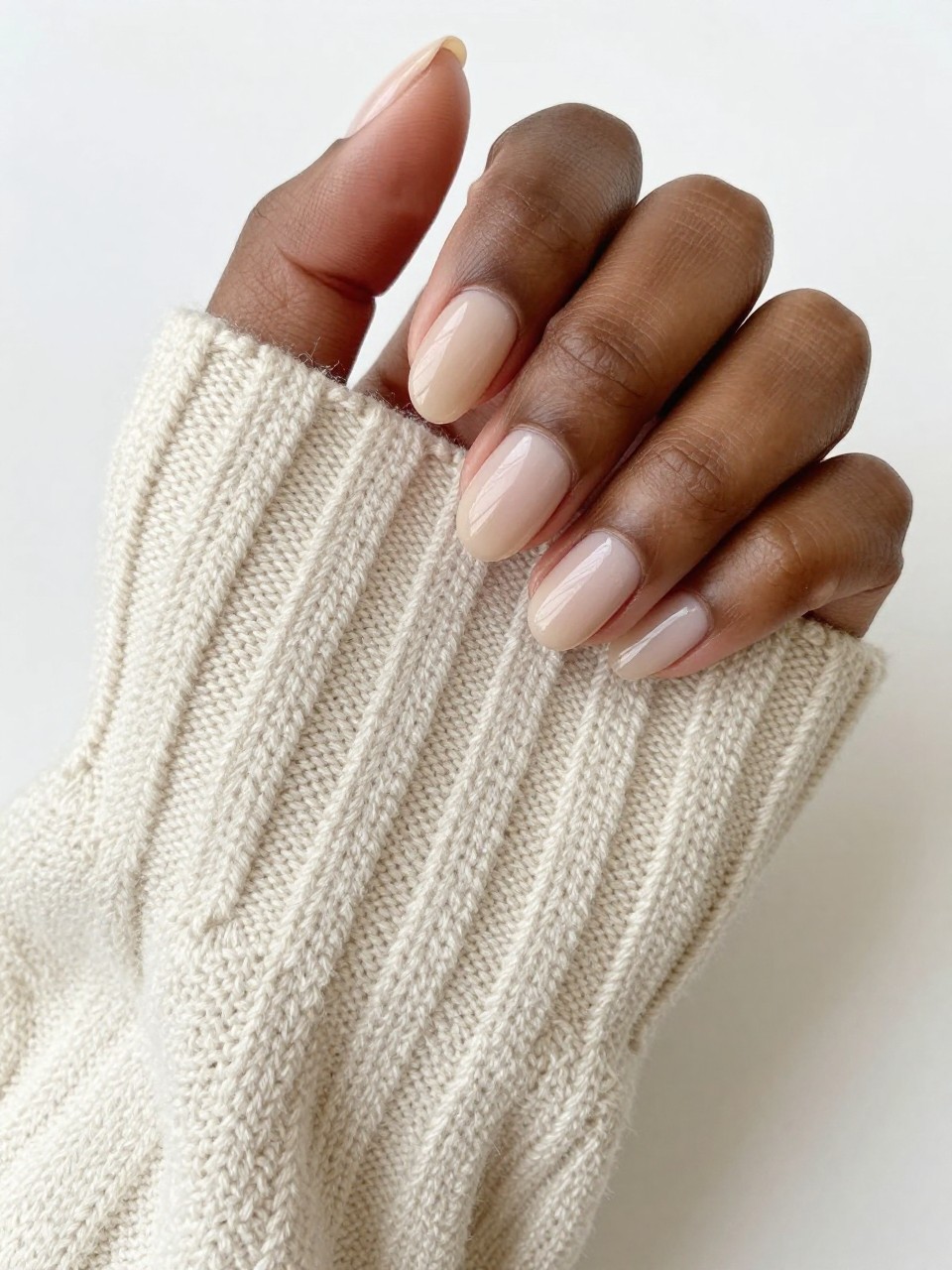Overhead shot of black feminine hands with very short, well-manicured nails resting on a creamy wool sweater. The nails are painted a transparent, matte beige that closely matches the natural nail bed. The look is pristine, soft, and focuses on perfect grooming.