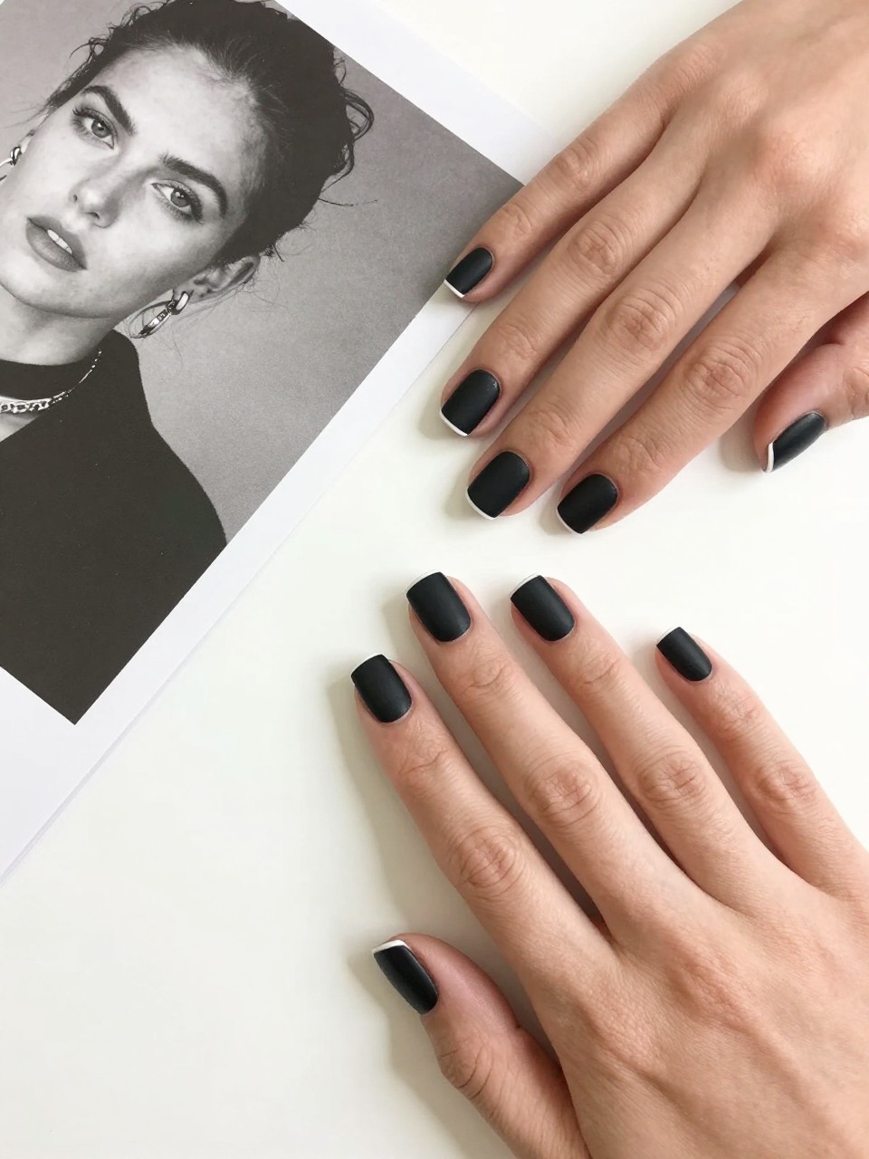 Top-down view of feminine hands with very short, squared-off nails placed next to a black and white photograph. The nails are a solid matte black. A hair-thin, perfect line of white polish edges the tip of each nail. The setting is a minimalist desktop with clean, bright lighting.