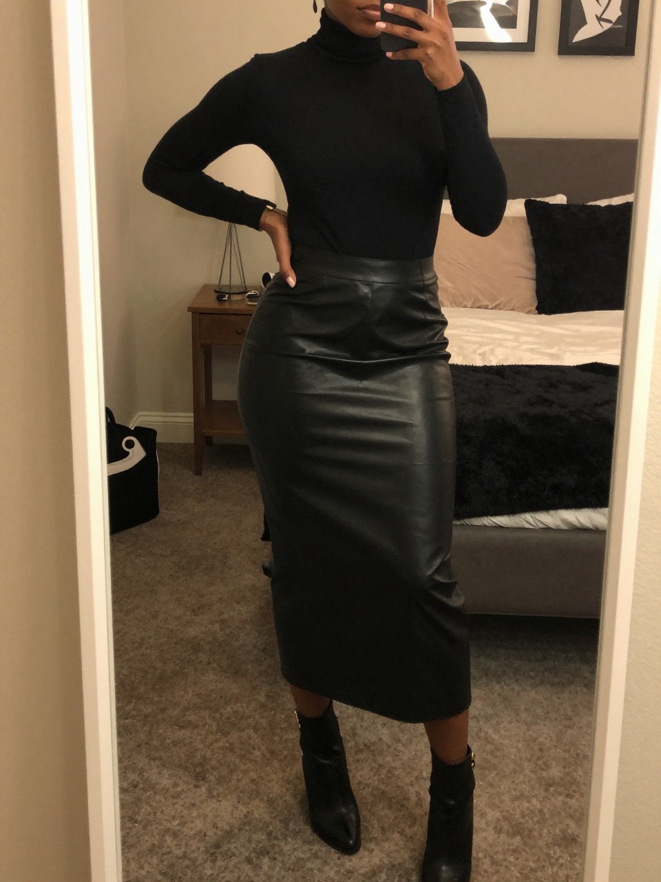 mirror selfie of a black woman in a leather long skirt and fitted turtleneck with ankle boots, standing with hand on hip, cropped at NECK so face doesn't show, in a feminine-themed bedroom, warm, moody lighting, iPhone photo quality.