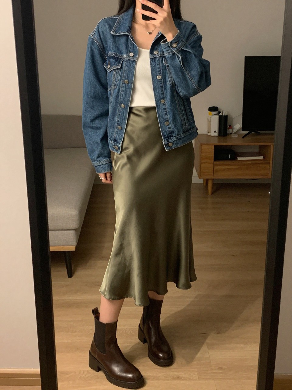 iPhone mirror selfie of a woman wearing a blue denim jacket over a white tank top with an olive green satin midi skirt and brown chunky ankle boots, one hand in her jacket pocket, cropped at NECK so face doesn't show, in a city apartment, warm moody lighting, iPhone photo quality.