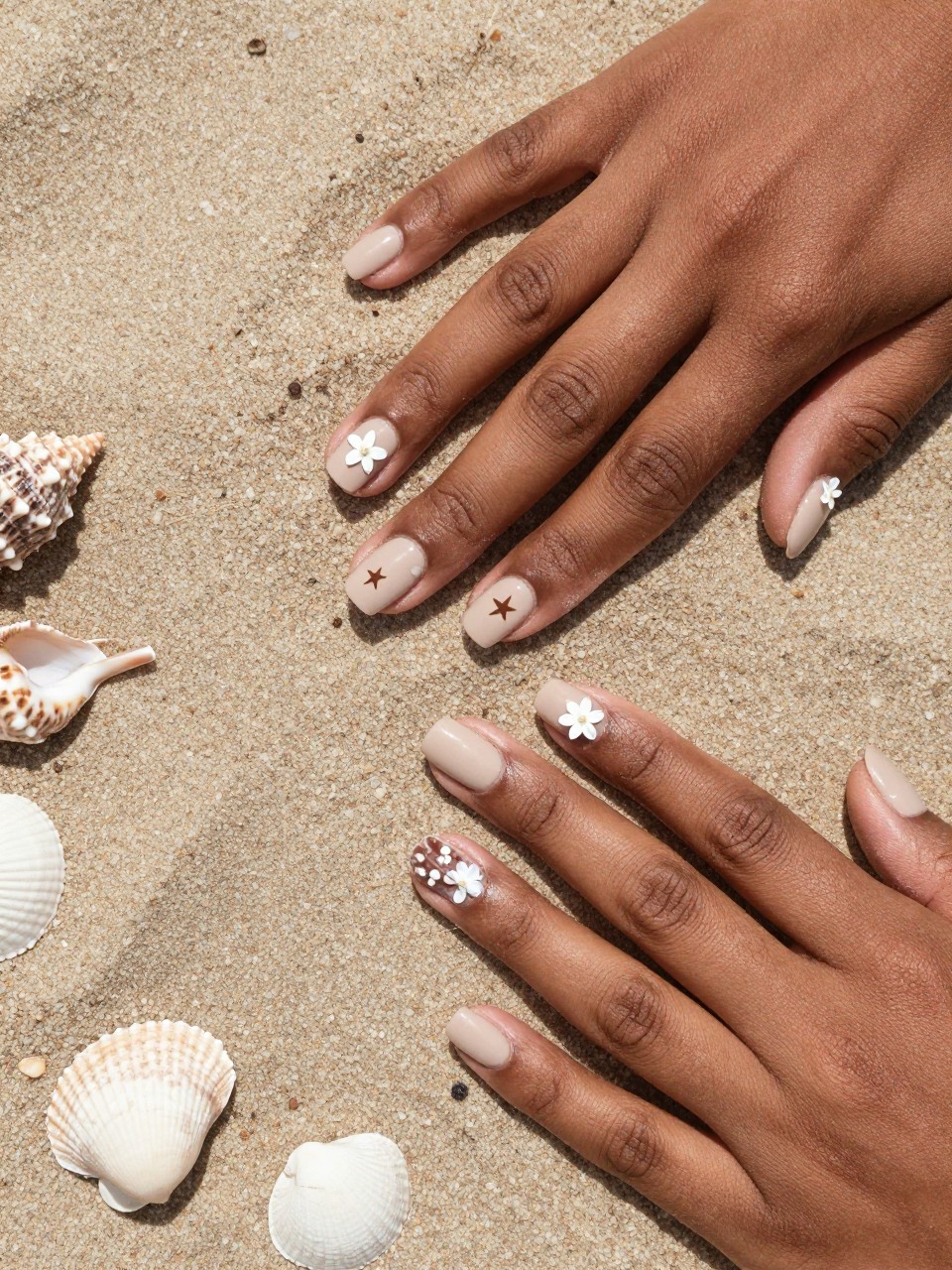Overhead shot of black feminine hands with rounded nails resting on actual sand and seashells. The nails are a matte sand beige. Tiny white starflowers and simple brown shell silhouettes are scattered on each nail. The light is bright and beachy.