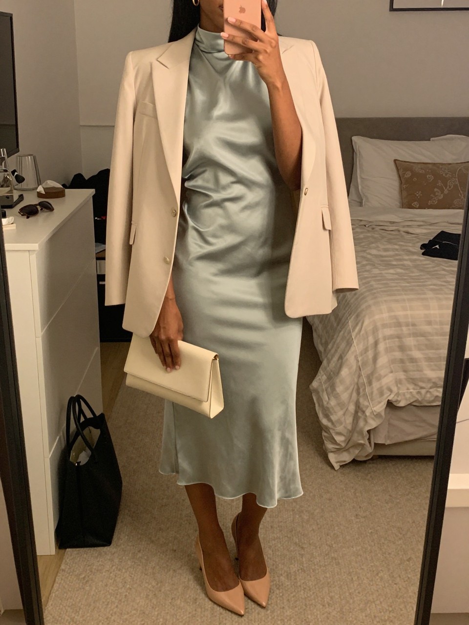 mirror selfie of a black woman wearing a soft sage satin high-neck midi dress with a cream light blazer, nude slingback pumps, an ivory structured clutch, and small gold hoop earrings, holding a phone in front of a mirror, cropped at NECK so face doesn't show, with a natural casual selfie pose, in a feminine-themed bedroom, warm moody lighting, including small lived-in details like a tote bag on the floor, sunglasses on a dresser, or soft bedding in the background, latest iPhone photo quality.