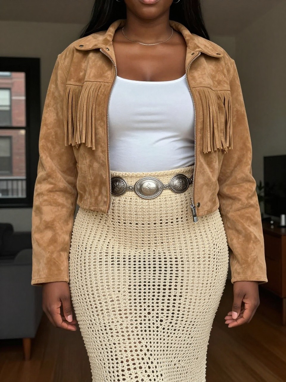 photo of a curvy black woman wearing a tan suede fringe jacket over a white tank top with a natural crochet midi skirt and a silver concho belt, one hand in her jacket pocket, cropped at NECK so face doesn't show, in a city apartment, warm moody lighting, iPhone photo quality.