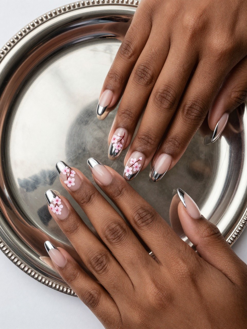 Bird's-eye view of black feminine hands with long nails next to a silver jewelry tray. The nails have mirror-like silver French tips. On the nails, there is a wispy cherry blossom branch with pink flowers is painted on the sheer nude base. The lighting highlights the chrome.